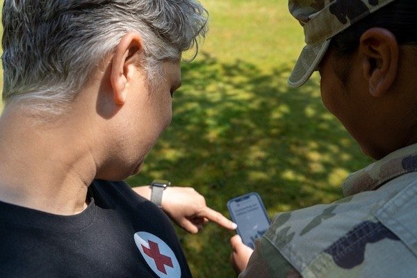 A Red Cross volunteer poses showing something to a uniformed servicemember on a mobile phone.