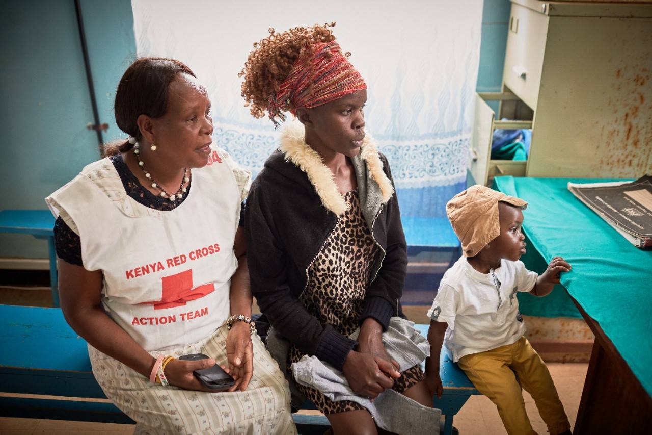 A mother, child and Kenyan Red Cross Society volunteer sit together in a clinic, waiting for an immunization.