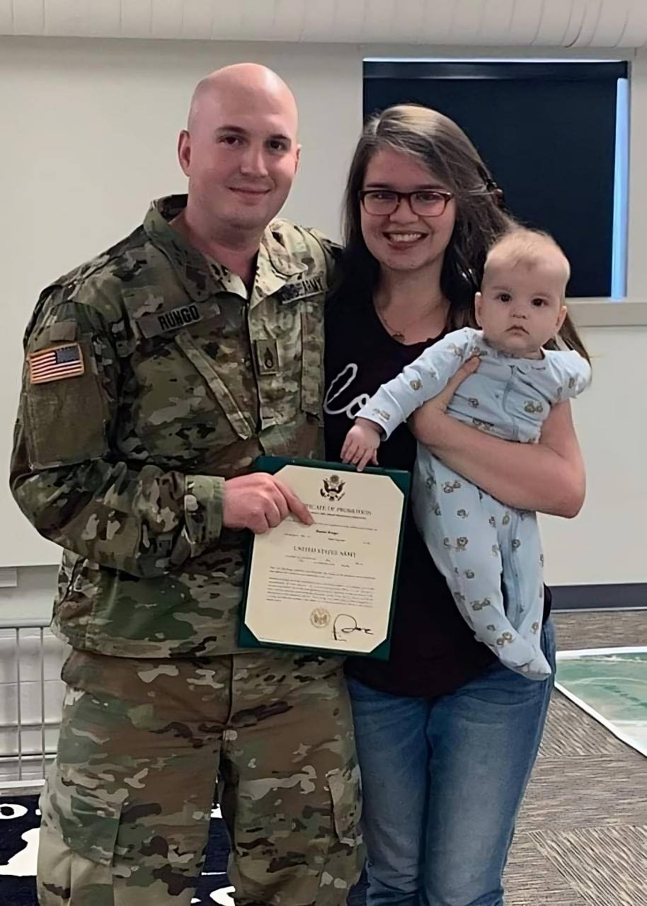 Un hombre con uniforme del Ejército mostrando su certificado de alistamiento mientras sonríe y abraza a su esposa. Ella tiene a su bebé en brazos.